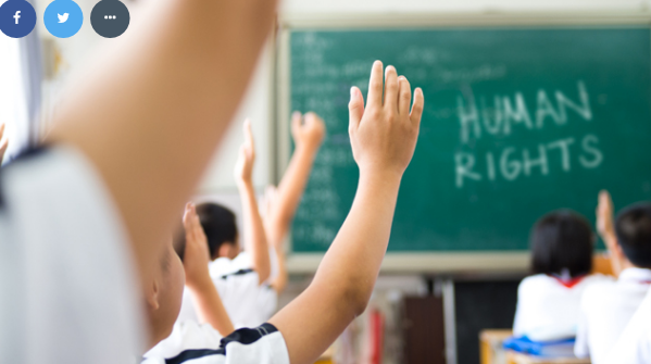 A classroom of children learning about human rights, each with their hand raised